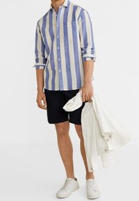 Homme portant une chemise à rayures verticales bleues et blanches, un short noir, des baskets blanches, tenant une veste blanche dans une main.