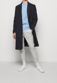 Soprabito scuro in misto lana con design a doppiopetto, felpa azzurro chiaro, chinos beige e sneakers bianche. Tessuto uniforme in tutta la superficie.