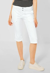 Bílé capri kalhoty se zapínáním na dva knoflíky, předními kapsami a ohrnutými lemy, doplněné o světlé tenisky a tričko s černými pruhy.