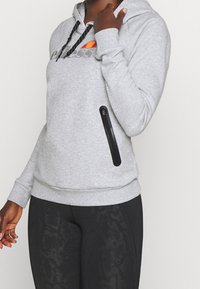 Haut à capuche gris avec une poche avant, capuche à cordon, et logo. Présente une poche latérale zippée avec une fermeture éclair noire et des poignets côtelés.