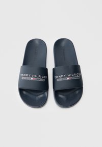 Tumši zili gumijas slaidu stila sandales ar platu siksnu, uz kuras izcelts zīmola nosaukums un dibināšanas gads kontrastējošā baltā un sarkanā krāsā.