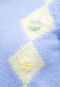 Ανοιχτό μπλε πλεκτό ύφασμα με κρεμ διαμαντένια σχέδια, το καθένα φιλοξενεί ένα χνουδωτό παστέλ λουλούδι από νήμα σε πράσινο, μωβ και κίτρινο χρώμα.