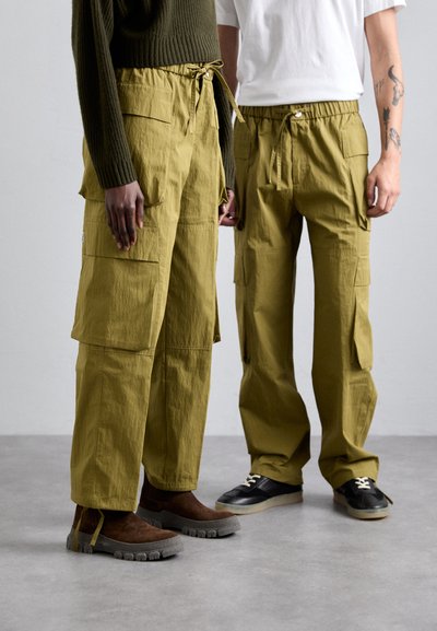 Olive cargo pants with multiple pockets and adjustable ties at the waist, paired with brown boots and black sneakers on a gray floor.