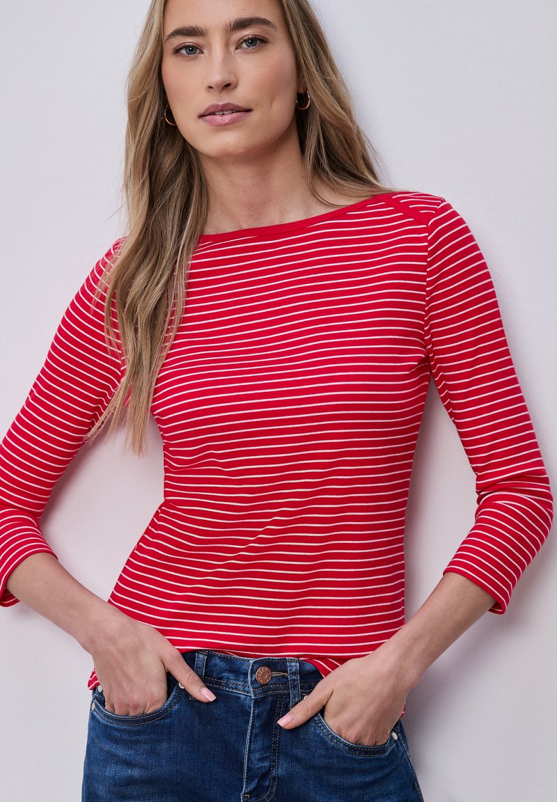 Femme aux longs cheveux blonds portant un haut à manches longues rayé rouge et blanc et un jean bleu, les mains dans les poches, devant un fond uni.