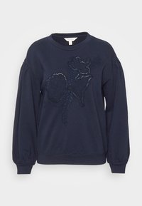 Felpa blu navy con maniche lunghe, polsini a coste e design floreale ricamato in tonalità scure sulla parte frontale. Tessuto morbido e texturizzato.