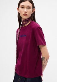 Burgunderfarget bomulls T-skjorte med korte ermer, med blå broderte "Calvin Klein"-logo og et mønstret halsbånd i nyanser av lilla og gull.