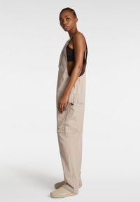 Beige Arbeitsoveralls mit offenen Seiten, verstellbaren Trägern und einer Brusttasche. Hergestellt aus leichtem Stoff mit glatter Textur.