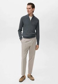 Grauer Reißverschluss-Pullover, beige Hose und hellbraune Schuhe. Der Pullover hat eine gerippte Textur; die Hose hat ein sauberes Design mit Bügelfalte.