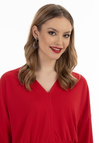 Piros V nyakú ruha hosszú ujjal; sima anyag; a modellen díszes ezüst és fekete fülbevalók vannak; hosszú, hullámos szőke haj.
