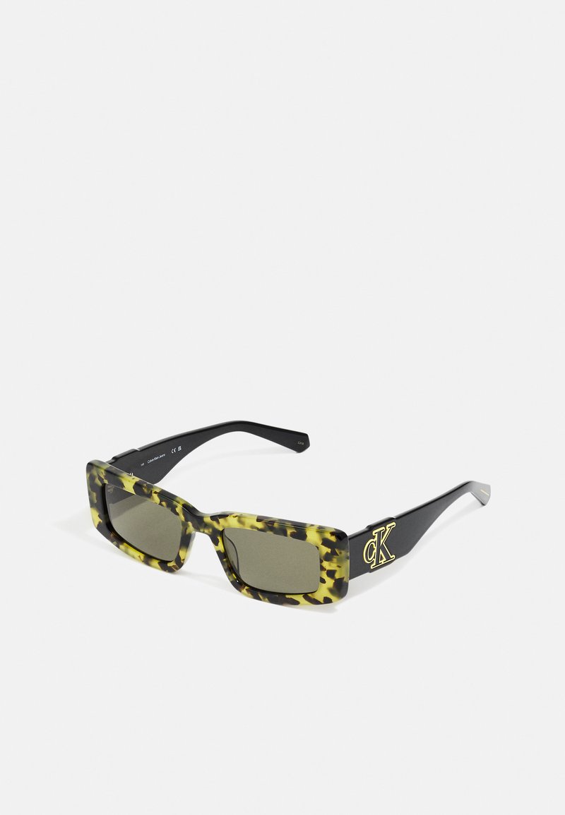 Suojalasit suorakaiteen muotoisena, joissa on keltainen ja vihreä simpukankuorikuosi, mustat varret sekä kultainen "CK" -logotieto temppeleissä.