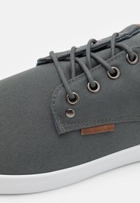 Szürke vászon sneaker fém fűzőlyukakkal, azonos színű fűzőkkel és barna bőr részlettel a fűzőlyukak közelében. Lapos fehér gumitalp.