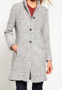 Grijze textuurjas met een kraag, voorzien van vijf zwarte knopen, zijzakken en een licht uitlopende zoom. De stof heeft een geweven patroon.