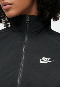 Μαύρο μπουφάν με φερμουάρ, κατασκευασμένο από υφή υφάσματος, με ψηλό γιακά, λείο φερμουάρ και λευκό λογότυπο Nike στο αριστερό στήθος.