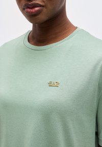 Κοντινό πλάνο ενός ατόμου που φοράει μια ανοιχτοπράσινη μπλούζα λαιμόκοψης με μικρό χρυσό λογότυπο EA7 Emporio Armani στο στήθος.