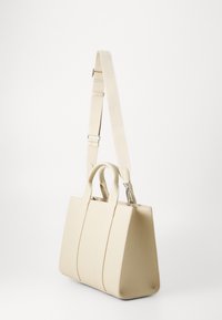 Sac à main beige avec une surface texturée, deux poignées supérieures et une sangle amovible. Présente un design structuré et angulaire avec des accents en métal argenté.