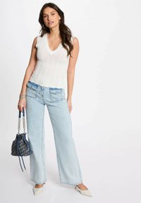 Top in maglia bianca a scollo a V con motivo a zigzag, abbinato a jeans a gamba larga blu chiaro. Gli accessori includono una borsa bucket blu navy e scarpe bianche a punta.