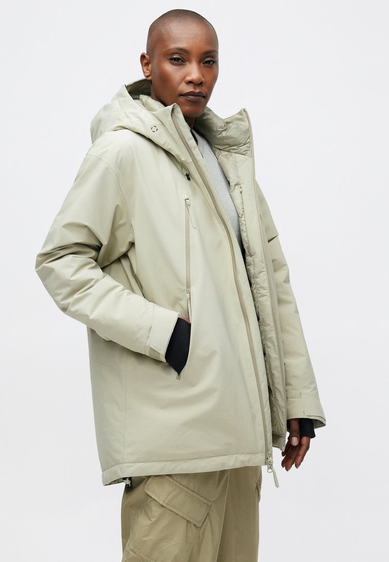 Helle graue Daunenjacke mit Kapuze, ausgestattet mit Reißverschlusstaschen und einer lockeren Passform. Glattes, glänzendes Äußeres mit elastischen Manschetten für zusätzliche Wärme.