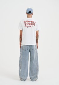 Bílá bavlněná trička s červeným grafickým textem na zadní straně, v kombinaci s světle modrými oversized džíny s květinovými vzory po stranách.