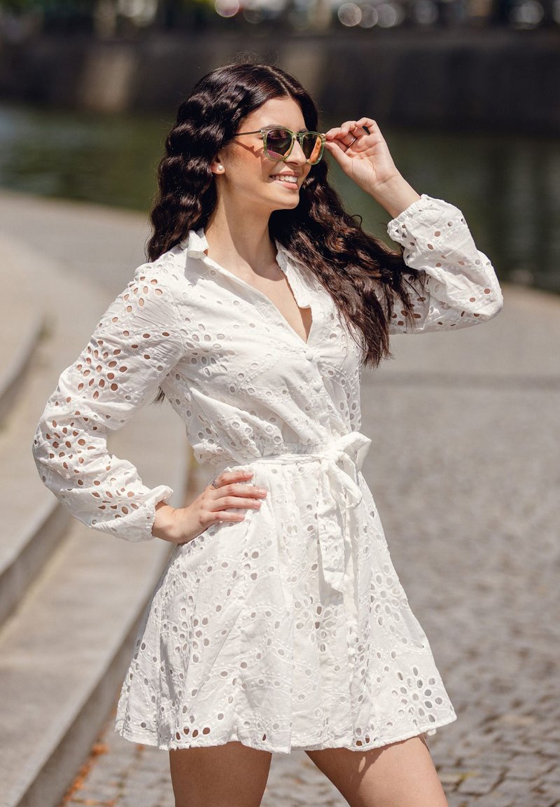 Bílá vyšívaná šaty s výřezovými vzory, dlouhé rukávy a zavázaný pas. Nosené s reflexními slunečními brýlemi. Venkovní prostředí u vody.
