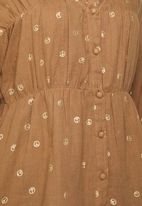 Blusa de algodão castanha com pequenos padrões de sinais de paz em ouro, frente com botões, cintura franzida e tecido texturizado.