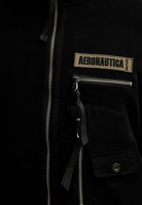 Giacca nera con zip in tessuto testurizzato, tasca frontale con zip, etichetta in tessuto che recita "AERONAUTICA" e tasca con bottoni a pressione.