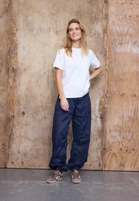 Moteris su ilgais blondiniais plaukais, dėvinti baltą trumpomis rankovėmis marškinėlį, laisvas tamsiai mėlynas kelnes ir rudus sportinius batelius, stovi prie medinės sienos ir šypsosi.