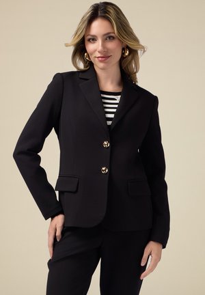 Blazer nero su misura con colletto a revers, bottoni dorati e due tasche frontali. Indossato sopra una maglia a righe bianche e nere.