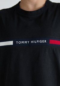 Crna pamučna majica s horizontalnim pojasom tekstom "TOMMY HILFIGER" u bijeloj, tamnoplavoj i crvenoj boji preko prsa.