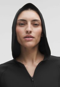 Îmbrăcăminte cu glugă neagră, cu fermoar, prezintă o textură netedă, o potrivire strânsă și un design minimal. Accent pe detaliile glugii și fermoarului.