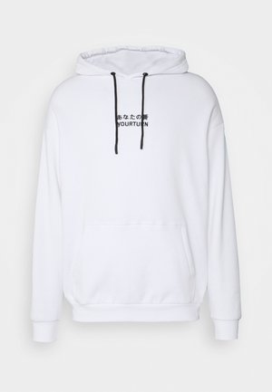 Hombre con sudadera blanca con cordones negros y texto, vaqueros azul oscuro con ligero desgaste y zapatillas negras con suelas blancas.