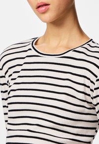 Približen posnetek osebe, ki nosi belo rebrasto srajco s črnimi vodoravnimi črtami in zaobljenim izrezom na vratu.