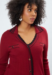 Roter gerippter Cardigan mit schwarzen Akzenten, gewelltem Ausschnitt, goldenen Knöpfen und zwei Brusttaschen, mit maßgeschneidertem Kragen und taillierten Ärmeln.