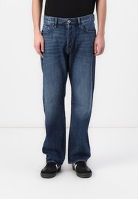 Jeans de mezclilla azul con un diseño de pierna recta, cinco bolsillos y cierre de botón. Llevados con zapatillas negras.