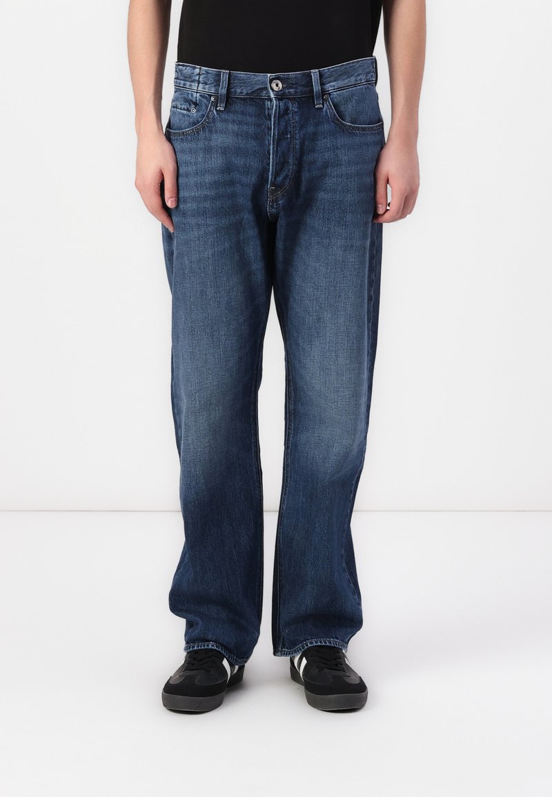 Jeans de mezclilla azul con un diseño de pierna recta, cinco bolsillos y cierre de botón. Llevados con zapatillas negras.
