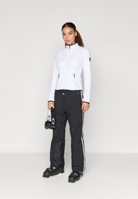 Valge tõmbluku järgi kinnitatav jakk kõrge kaelusega, millele on sobitatud mustad suusapüksid, millel on valged küljeribad. Mustad suusajalatsid ja rihmaga kott on käes.