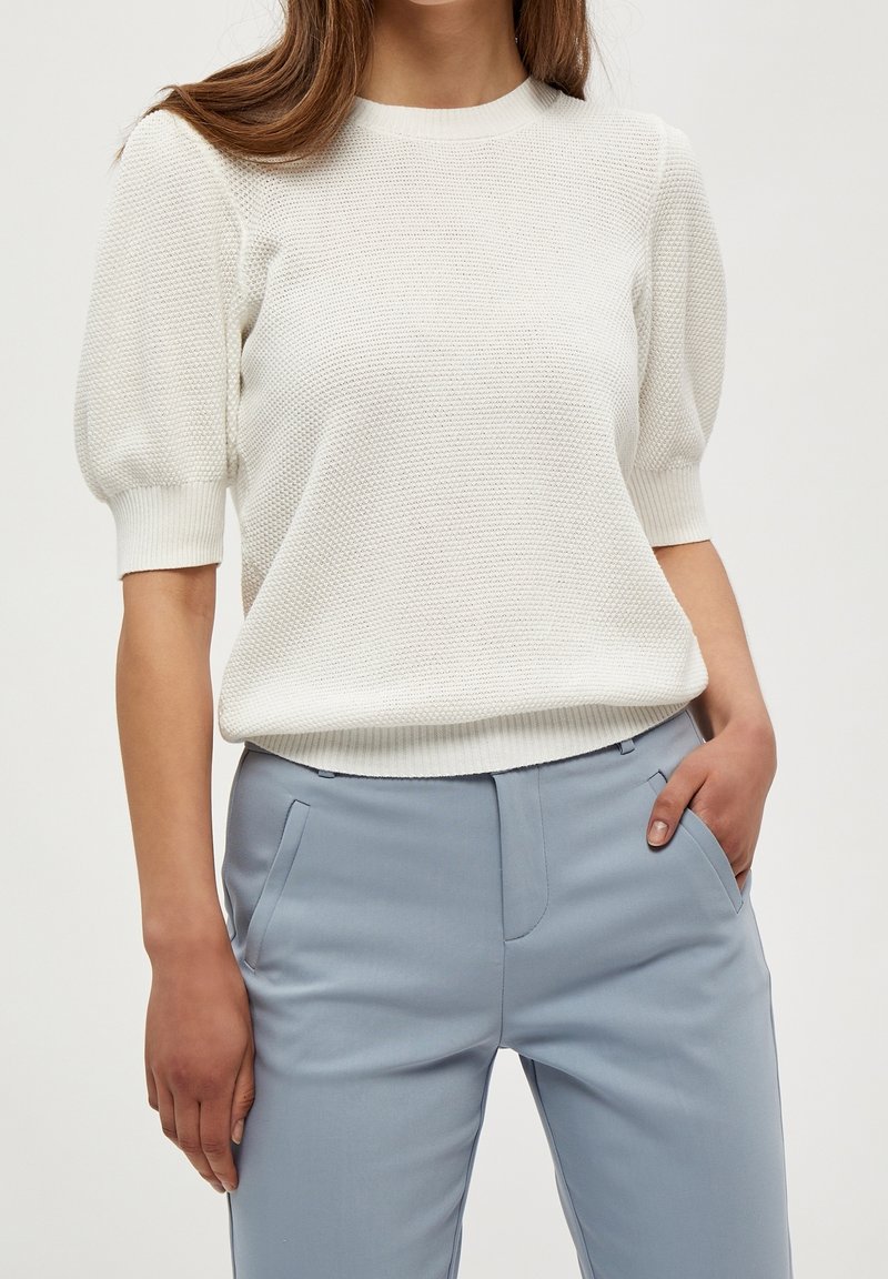 Pull en tricot blanc avec des manches courtes bouffantes et un col rond, associé à un pantalon ajusté bleu clair. Texture douce et coupe décontractée.
