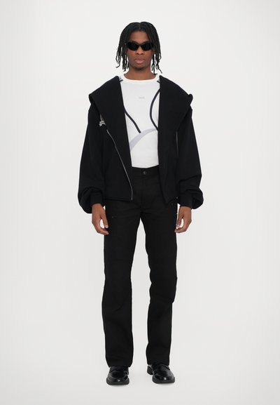 Jachetă oversize neagră cu glugă și fermoar frontal, cămașă albă cu design gri, pantaloni negri și încălțăminte neagră, accesorizată cu ochelari de soare.