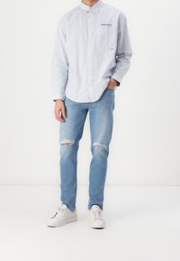 Jasnoniebieska koszula na guziki w paski z białymi guzikami, zestawiona z przetartymi niebieskimi dżinsami i białymi trampkami. Logo Tommy Jeans na kieszeni koszuli.