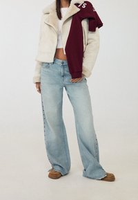 Krátká bunda z krémového umělého kožíšku se zipem, světle modré džíny s širokými nohavicemi a hnědé mokasíny. Maroon mikina přehozená přes rameno.