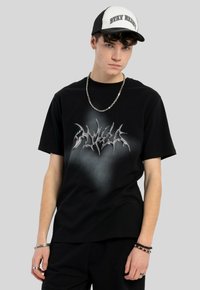 Czarny t-shirt z krótkim rękawem z metalicznym wzorem graficznym z przodu. Model nosi czapkę w czarno-białym kolorze, naszyjnik z łańcuchem i bransoletki.