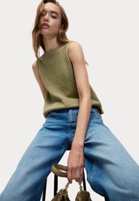 Olijfgroene gebreide tanktop met een gestructureerde afwerking, gecombineerd met lichtblauwe wijde jeans. Het model houdt een beige handtas vast.