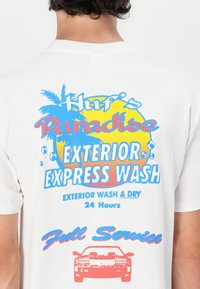 Biała bawełniana koszulka z kolorowym tekstem graficznym przedstawiającym palmy, słońce i ilustrację samochodu, oznaczona napisem "Huf's Paradise Exterior Express Wash".
