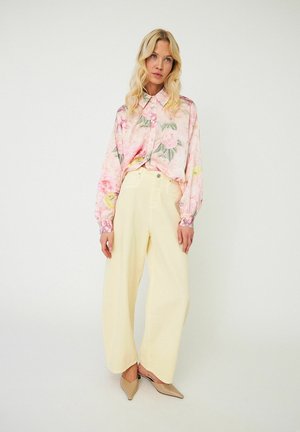 Blonde woman wearing a pink floral blouse, wide pale yellow pants, and beige pointed heels, standing against a plain white background.