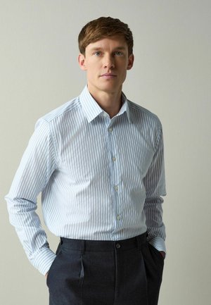 Mann mit kurzem braunem Haar, der ein hellblau gestreiftes Hemd mit Knöpfen und eine dunkle Hose trägt. Er steht mit den Händen in den Taschen vor einer schlichten Wand.