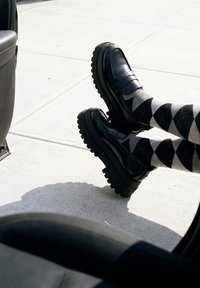 Jambes portant des mocassins noirs massifs et des chaussettes losanges noires, grises et blanches, étendues à l'extérieur d'une portière de voiture sur un trottoir en béton.