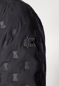 Black jacket with a textured surface featuring a geometric pattern. Includes a rubber brand patch on the left sleeve.