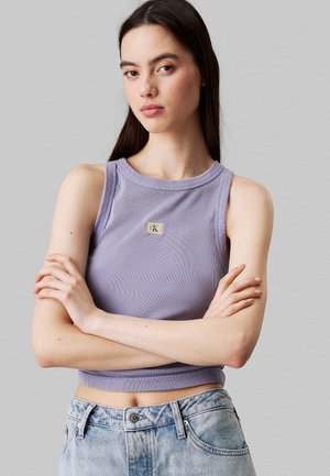 Lavendel geribbelde crop top met een ronde halslijn, voorzien van een klein gouden embleem op de borst. Model draagt lichtblauwe high-waisted jeans.