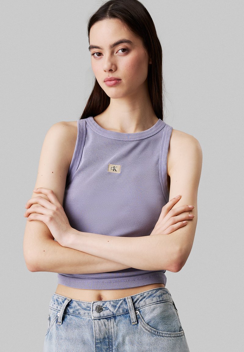Top corto a coste lavanda con scollatura rotonda, dotato di un piccolo emblema dorato sul petto. Il modello indossa jeans azzurri a vita alta.