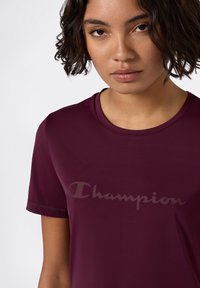 Mujer joven con pelo corto y rizado, vistiendo una camiseta Champion color burdeos con un logo sutil en el pecho, contra un fondo claro y liso.