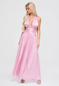 Femme blonde portant une robe rose fluide à col halter avec un décolleté profond et des talons argentés ouverts sur un fond clair uni.